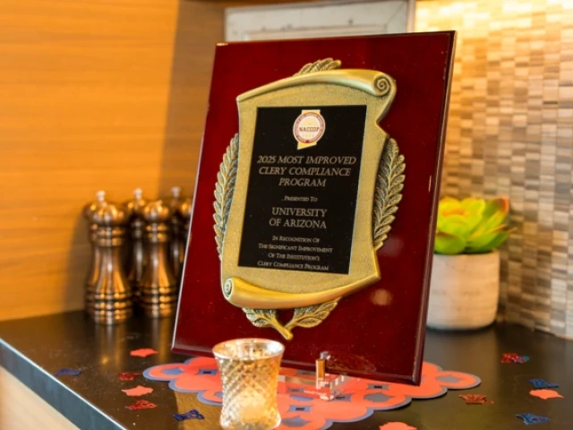 A close-up of a plaque reading "2025 Most Improved Clery Compliance Program, presented to University of Arizona," displayed on a countertop with small decorative items around it.