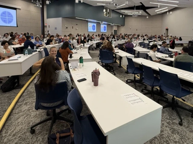 Attendees fill a large lecture hall during the July 30 Conflict Resolution and De-escalation Skills Symposium at the University of Arizona. Participants sit in groups at long white tables, engaging in discussion during an interactive portion of the event. Multiple presentation screens display a slide with the word “practice” and a blue circular graphic divided into four sections.