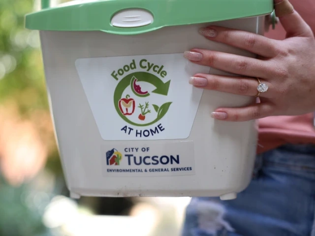 A close-up of a person holding a small white composting bin labeled “Food Cycle at Home,” with logos for the City of Tucson Environmental & General Services. The person’s hand, wearing a large oval-cut engagement ring, rests on the bin.