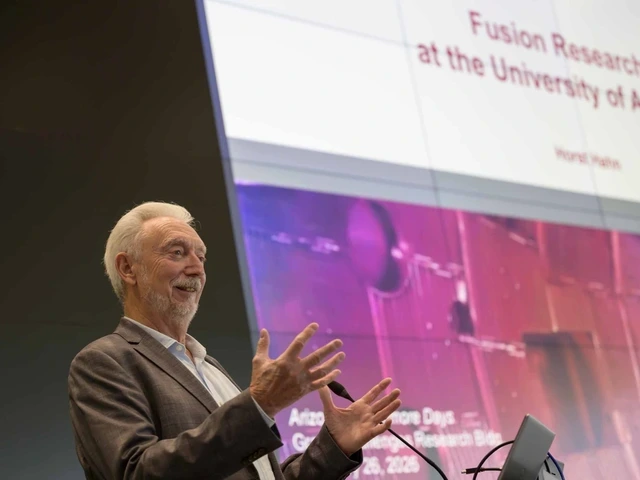 A man in a brown suit stands in front of a podium with his hands in front of him. Behind him is a screen with a presentation that reads "Fusion Research a the University of Arizona"
