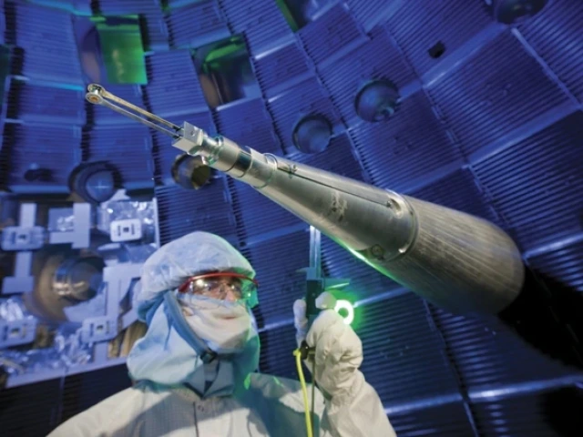 A technician in full cleanroom attire, including a face shield and hood, stands near a long, conical instrument used to position fuel targets inside the National Ignition Facility. The background shows the interior of the target chamber, with glowing blue lighting and metal structures creating a high-tech atmosphere.