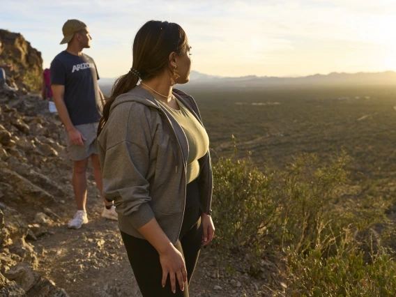 Two hikers, a man and a woman, pause along a rocky trail during sunset, overlooking a vast desert landscape. The woman, in the foreground, looks off into the distance while wearing a light gray hoodie and dark leggings. The man, in the background, wears a University of Arizona t-shirt and a baseball cap, standing slightly behind her. The warm, golden light of the setting sun illuminates the scene, casting long shadows across the desert terrain.