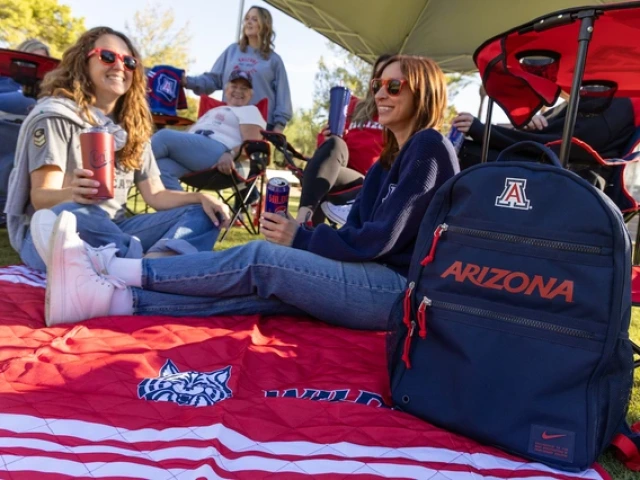 People relax under a canopy at a tailgate on a sunny day, sitting on a red University of Arizona blanket. A navy backpack with “Arizona” and the “A” logo is in the foreground.