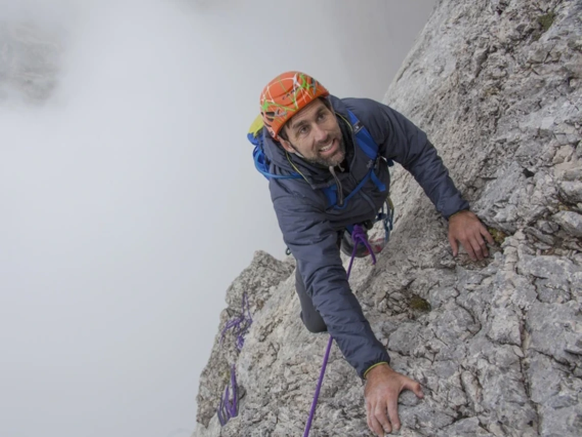 A climber in a gray jacket and bright orange helmet ascends a steep, rugged rock face using ropes and climbing gear. He looks up with determination, gripping the stone firmly as mist swirls around the mountain behind him.