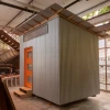 an 8-by-12-foot shelter with metal siding and an orange door sitting inside a large manufacturing shop