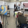 University of Arizona art students paint at easels in a studio while a string quartet performs live music in the background. The students are focused on their colorful abstract paintings, with palettes and paint supplies on nearby tables. The musicians, standing in front of a red tapestry and dress forms, include three violinists and a cellist.