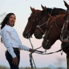 A woman with long dark hair wearing dark denim jeans and a long sleeved white formal shirt stands in front of four horses in an outdoor environment. The sun is setting in the background. The woman is holding leads to all four horses.