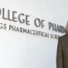 Jack R. Cole, wearing a brown plaid suit and patterned tie, stands smiling in front of a white wall with large lettering that reads "College of Pharmacy Skaggs Pharmaceutical Sciences."